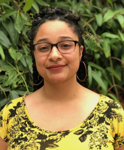 headshot photo of a Black woman smiling, wearing glasses, gold hoop earrings, a yellow dress with black flowers and leaves. She is standing in front of a green leafy bush